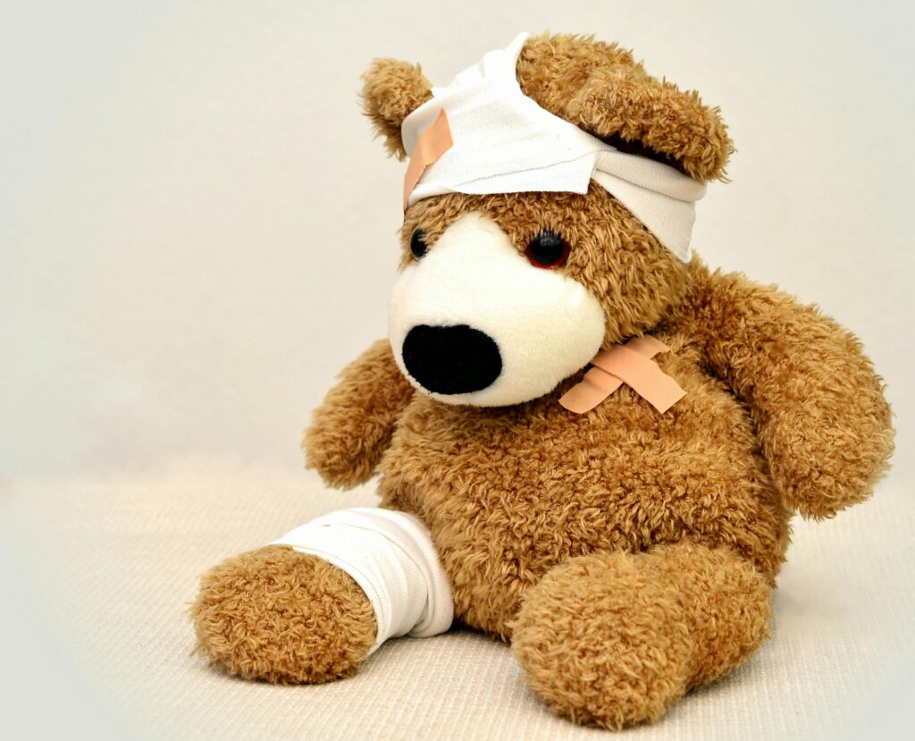 Cute brown teddy bear with bandages wrapped around its leg and head, with crisscrossed bandaids over the heart.